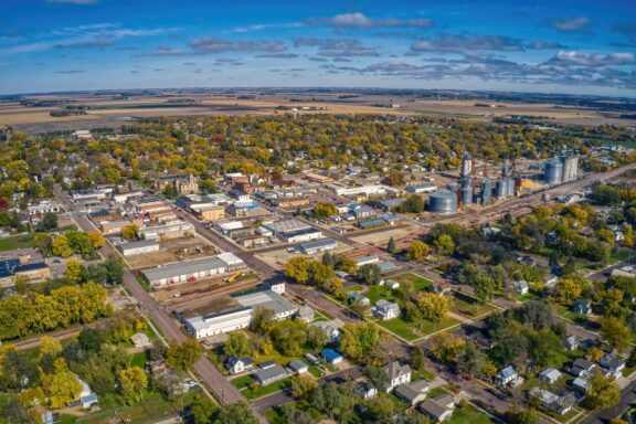 Aerial view of a small town with residential areas, commercial buildings, grain silos, and a railroad on a sunny day with scattered clouds.