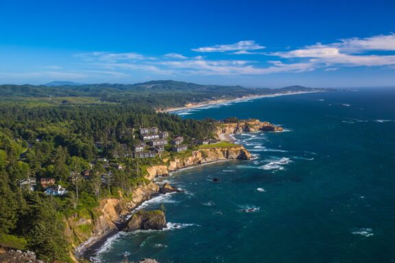Scenic Coastal View with Basalt Outcroppings and Lush Greenery Aerial view of a rugged coastline with a small community surrounded by forest, overlooking the Pacific Ocean, under a clear blue sky.