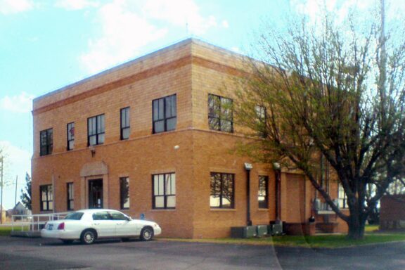 A two-story brick building on a sunny day with a white car parked in front. Trees with spring foliage are visible around the building.