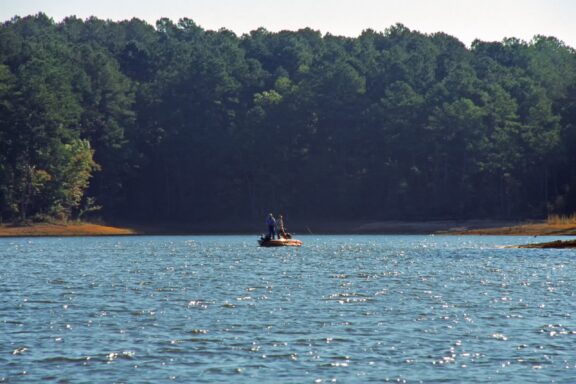 A small boat with two people fishing on a serene lake with a backdrop of dense, green forest under a clear blue sky.