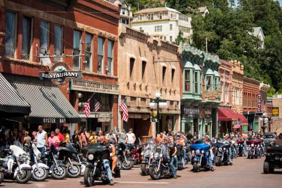 A bustling historic street lined with classic architecture, featuring a crowd of people with parked motorcycles, American flags, and old-fashioned storefronts under a clear sky.