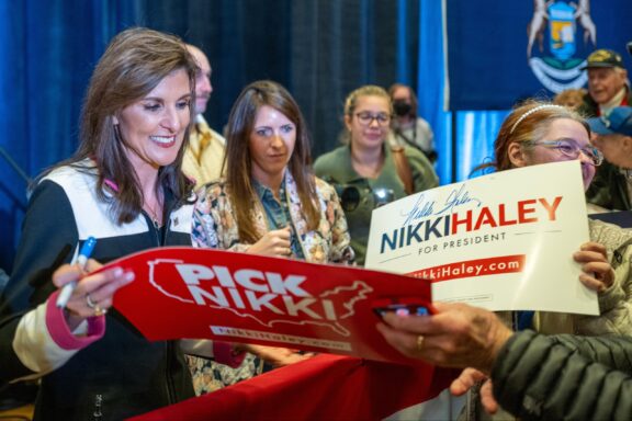 Nikki Hayley smiling and signing a red "PICK NIKKI" campaign sign for an eager crowd, with other supporters holding a "NIKKI HALEY FOR PRESIDENT" sign in the foreground.