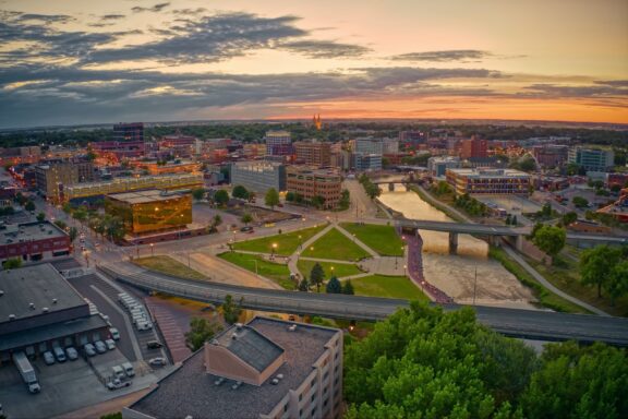 Aerial view of a city at dusk, featuring a river flowing through, multiple buildings with lights on, roads with sparse traffic, a bridge crossing the river, and a park with pathways and green spaces, all under a vibrant sunset sky.