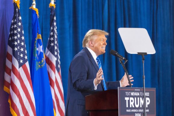 Donald Trump stands at a lectern with microphones, speaking and gesturing, with several American flags and a state flag in the background.