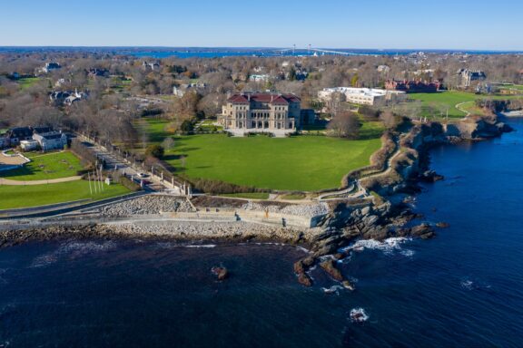 Aerial view of a grand historic mansion with a large green lawn, overlooking a rocky coastline with a clear blue sea, and a bridge in the distant background.