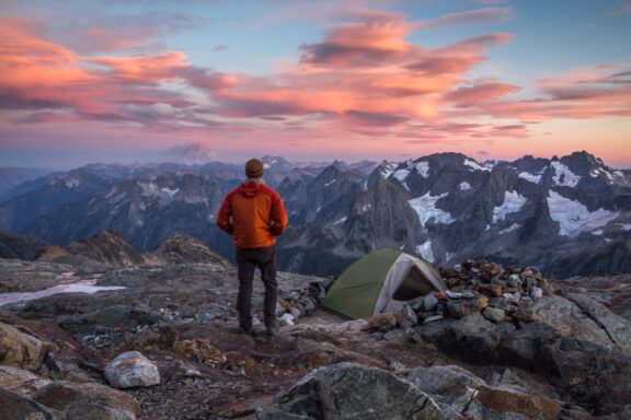 A person in an orange jacket and beanie standing on a rugged mountain overlook and gazing at a colorful sunset, with a green tent pitched nearby on rocky terrain.