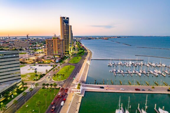 Aerial view of a coastal cityscape at dusk with a high-rise hotel, marina full of sailboats, a palm-lined boulevard, and a bridge in the background.