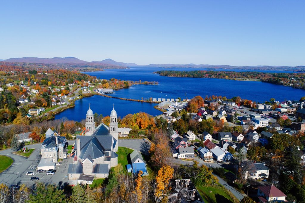 Aerial view of a coastal town with autumn-colored trees, featuring a prominent church, surrounding homes, and a marina leading out to a large lake with distant hills under a clear blue sky.