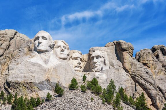 The Mount Rushmore National Memorial with sculptures of the heads of four United States presidents carved into the granite face of Mount Rushmore in the Black Hills region of South Dakota, under a blue sky with wispy clouds.