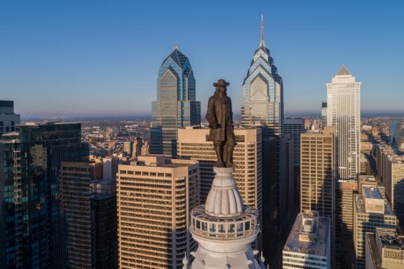 Aerial view of a city skyline with prominent skyscrapers, featuring a statue of William Penn in the foreground atop a building, under a clear blue sky.