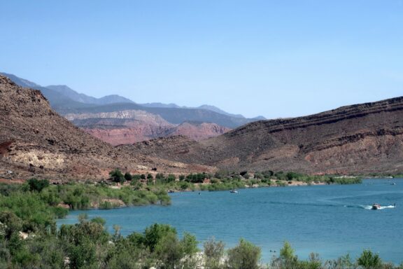 A serene lake with clear turquoise waters, flanked by rugged hills under a blue sky, with a motorboat cruising and distant red rock formations in the background.