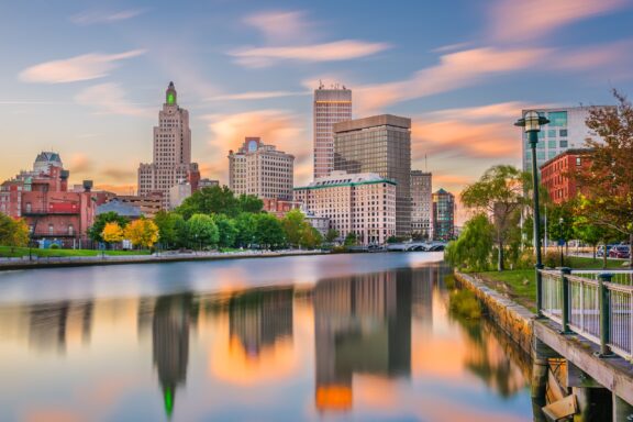 A city skyline at dusk with buildings reflected in the calm water of a river, under a colorful sky streaked with clouds.