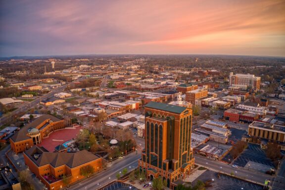 Aerial view of a city at sunset with a prominent brick tower in the foreground and a mix of commercial buildings and streets extending into the background under a pink and blue sky.