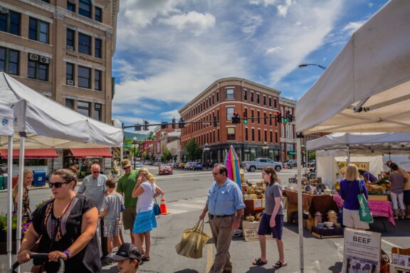 Street scene at an outdoor market with tents, vendors selling goods, and people browsing, set against a backdrop of historic city buildings and clear blue sky.