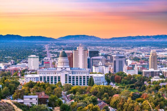 Aerial view of a city skyline at dusk with colorful sunset skies, prominent capitol building in the foreground, surrounded by various modern and traditional buildings, with a backdrop of distant mountains.