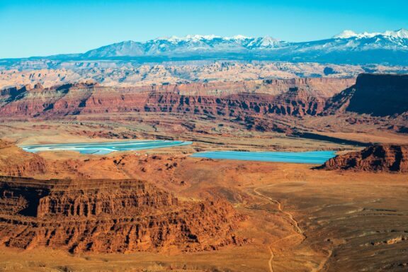 Aerial view of a winding river cutting through a red desert canyon with snow-capped mountains in the background.