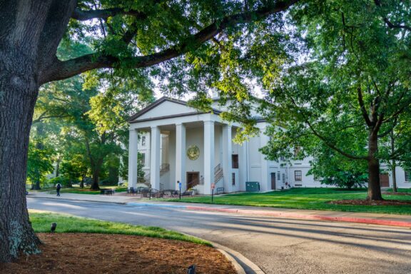 A stately white building with large columns and a central clock, surrounded by lush green trees, with a pedestrian walking along a pathway in the foreground.