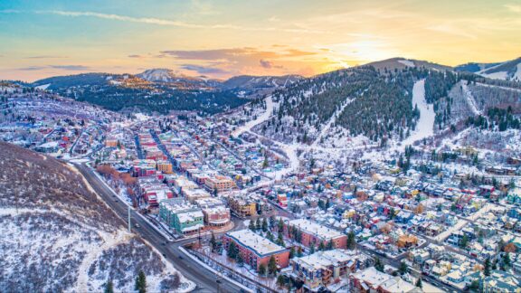 Aerial view of a colorful mountain town during winter with snow-covered rooftops and streets, surrounded by forested hills and a setting sun in the background.