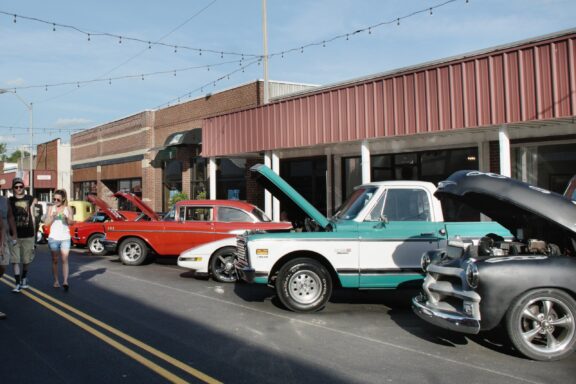 A classic car show on a sunny day with the hoods of various vintage cars open; people are walking and observing the cars in a street flanked by brick buildings with string lights above.