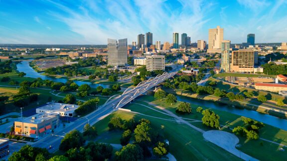 Aerial view of a cityscape with modern buildings, a meandering river, green parkland, and a distinctive curvy bridge under a clear blue sky.