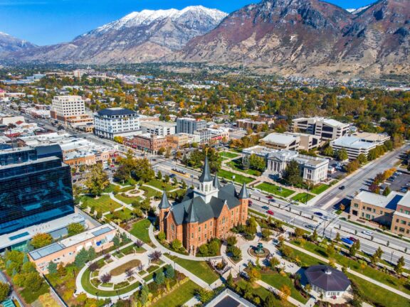 Aerial view of a downtown area with a mix of historic and modern buildings, landscaped gardens, roads with light traffic, and mountains in the background.