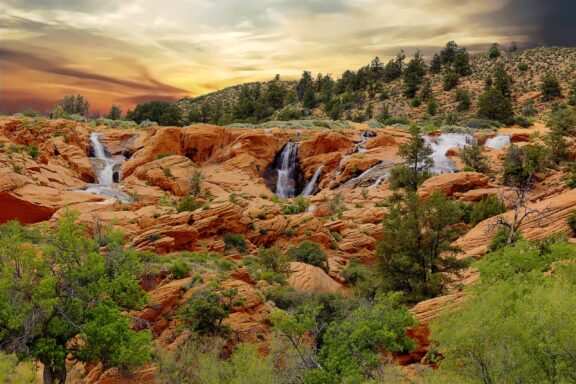 A scenic view of a small waterfall cascading over vibrant red rock formations surrounded by lush greenery with a dramatic sunset sky in the background.