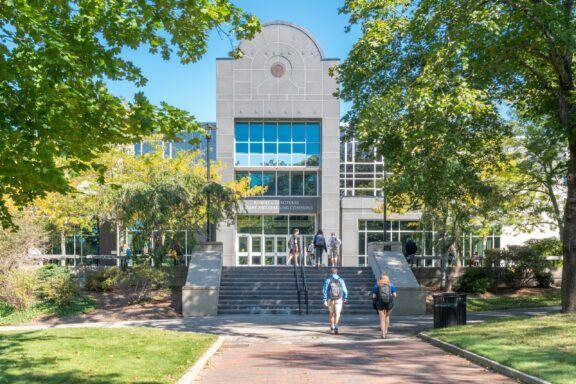 A modern building with a large glass facade and the text "Robert L. Carothers Library and Learning Commons" over the entrance. Students are walking up and down the stairs and along the brick walkway in front of the building, flanked by green trees and a clear sky.