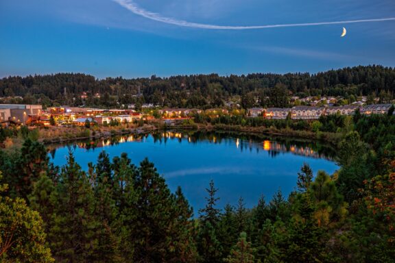 Twilight view of a calm lake reflecting the illuminated buildings and lights on the shore, with a crescent moon in the sky, surrounded by forested hills.