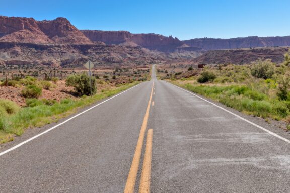 A straight two-lane road with yellow dividing lines leading towards distant red rock mesas and clear blue skies.