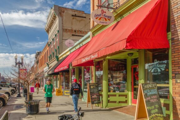 A vibrant street view with pedestrians walking past the Great Harvest Bread Co. with red awnings, under a clear blue sky.