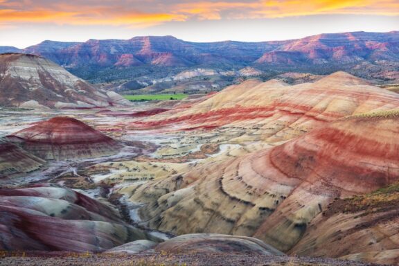 Colorful Strata of the Painted Hills at Sunset in John Day Fossil Beds National Monument, Wheeler County A vibrant landscape of hills with colorful stratified earth formations in red, yellow, and brown hues under a sunset sky with light clouds.