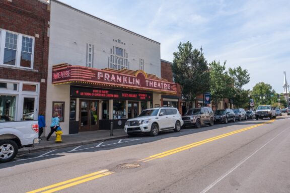 The Franklin Theatre with a marquee sign, located on a busy street lined with parked cars and a few pedestrians walking by.