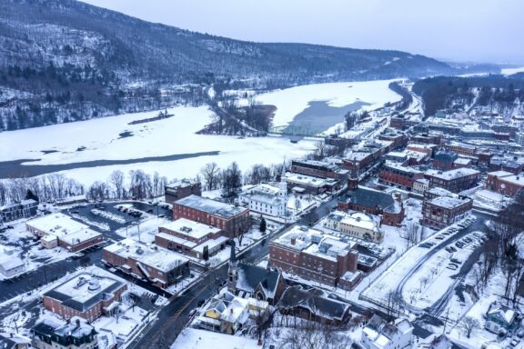 Aerial view of a snow-covered small town with buildings along riverbanks, a bridge crossing the partially frozen river, and snow-dusted hills in the background.