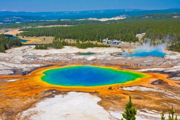 A vivid aerial view of the Grand Prismatic Spring in Yellowstone National Park with its bright colors ranging from blue to orange, and a forested background.