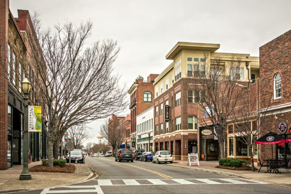 A typical urban street scene with a mix of brick and modern buildings, parked cars, pedestrian crossings, bare trees, and storefronts, under an overcast sky.