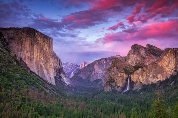 A sunset over Yosemite Valley with a colorful sky, featuring El Capitan on the left, Bridalveil Fall on the right, and Half Dome in the distance.