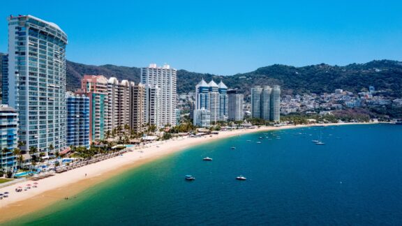 A panoramic view of the coastal cityscape of Acapulco featuring a sandy beach lined with palm trees and tall buildings, with boats dotting the blue waters and green hills in the background.