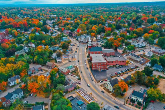 Aerial view of downtown Dover, New Hampshire with a mix of autumn-colored trees and traditional architecture, featuring a prominent clock tower near a crossroads, surrounded by dense woodland in the background.