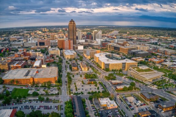 Aerial View of Des Moines, Iowa Skyline Aerial view of a cityscape at dusk, featuring a mix of high-rise and low-rise buildings, with the tallest skyscraper near the center surrounded by various commercial buildings, streets, parking lots, and patches of greenery, under a partly cloudy sky with a soft glow from the setting sun.