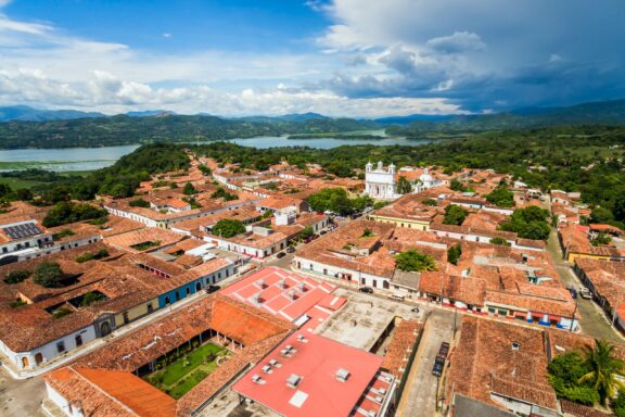 Aerial view of a historic colonial town with a white church, terracotta-roofed buildings, and streets, with a lake and mountains in the background under a cloudy blue sky.