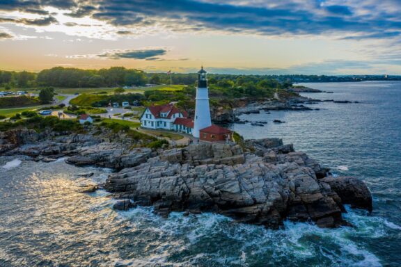 Fort Williams Park in Cape Elizabeth, Maine: Aerial view of a lighthouse on a rocky promontory near the coast at sunset with waves crashing on the shore, surrounded by greenery and a partially cloudy sky.