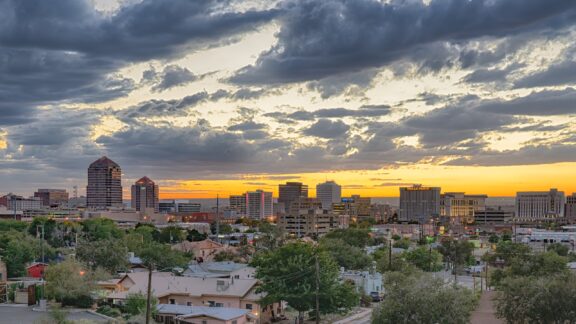 Sunset view over a city skyline with illuminated buildings under a dramatic cloudy sky.