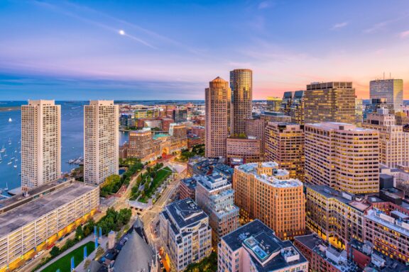 Aerial view of a densely populated city at dusk with skyscrapers, a waterfront, and a colorful sky.