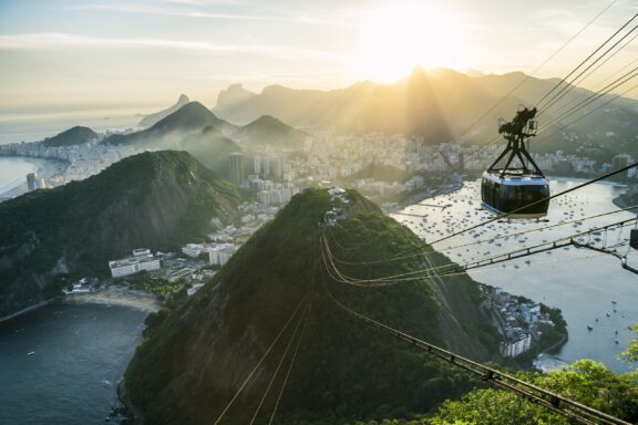 A cable car descending over a lush green hillside with a panoramic view of Rio de Janeiro's cityscape and coastline at sunset.