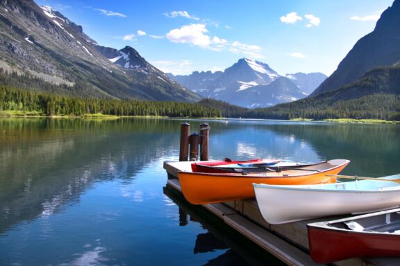 A serene mountain lake with colorful kayaks lined up on a wooden dock, with a backdrop of forested slopes and snow-capped mountain peaks under a clear blue sky.