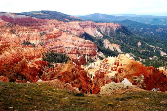 A panoramic view of Cedar Breaks National Monument showcasing the vibrant red and orange hues of the intricate rock formations and canyons with green forested slopes in the background under a partly cloudy sky.