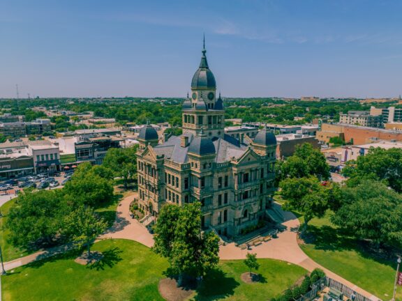 Aerial view of a historic courthouse with a domed clock tower, surrounded by well-manicured lawns, pathways, and a backdrop of a suburban town under a clear blue sky.