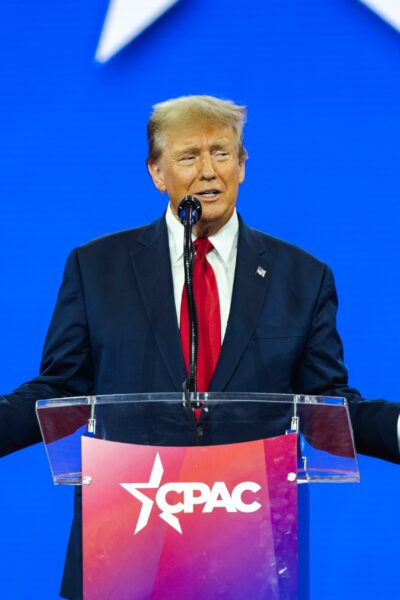 Donald Trump in a dark suit, white shirt, and red tie speaking at a lectern with the CPAC logo, against a blue background with white star graphics.