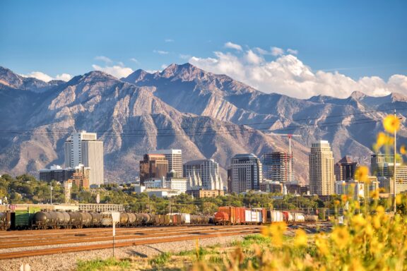 A city skyline with modern buildings in the foreground, a freight train in the midground, and a backdrop of towering mountains under a clear sky.