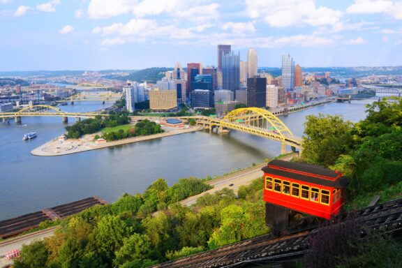 Aerial view of Pittsburgh's cityscape featuring multiple bridges over the Allegheny River, downtown skyscrapers, and the Duquesne Incline with a red cable car in the foreground.